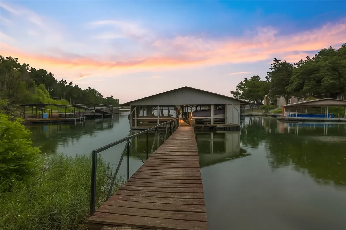 The image showcases a tranquil lakefront scene at sunset, with golden light reflecting off the calm water. A charming wooden dock extends from the shore, leading to a boathouse nestled along the water's edge. The setting invites relaxing by the water, boating, fishing, or simply soaking in the peaceful lake atmosphere.