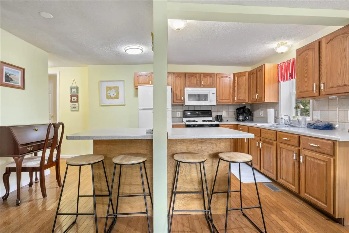 A wide view of the kitchen featuring a spacious island, wooden cabinets, and inviting bar stools. The dining area is visible nearby, with natural light filling the space and hints of the outdoors visible through windows.