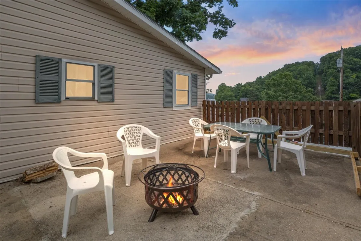 An exterior view of a fire pit area with comfortable outdoor seating and a table, set against a backdrop of natural scenery. The setting is warm and welcoming, ideal for socializing, dining, or enjoying the starry night sky.