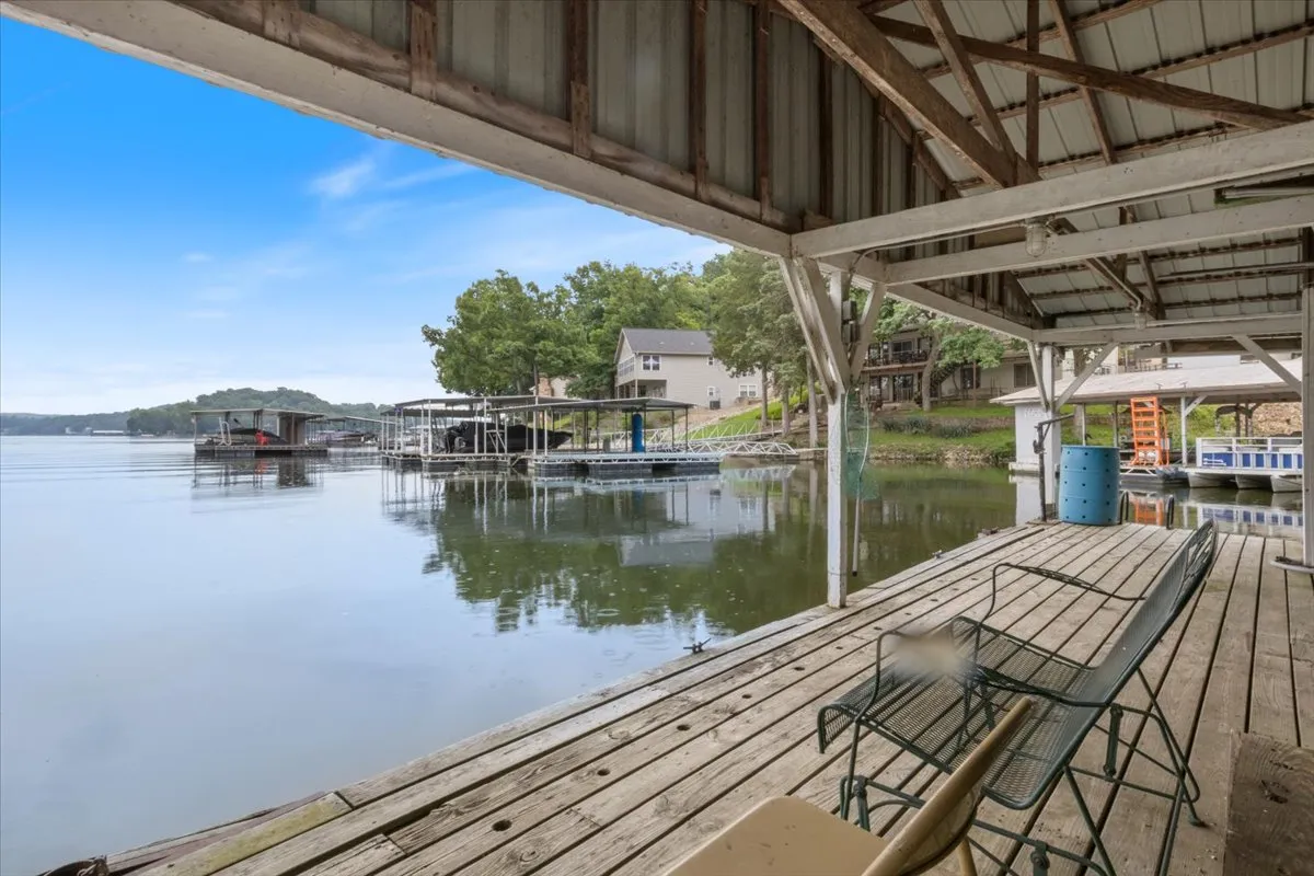 A wide shot of the lakefront showing a wooden dock with boats moored, surrounded by lush greenery. The calm water stretches to the horizon, reflecting the colors of the sky, with a hint of sunset and a peaceful, inviting atmosphere. The dock is clearly accessible, ideal for fishing, boating, or simply relaxing by the water.