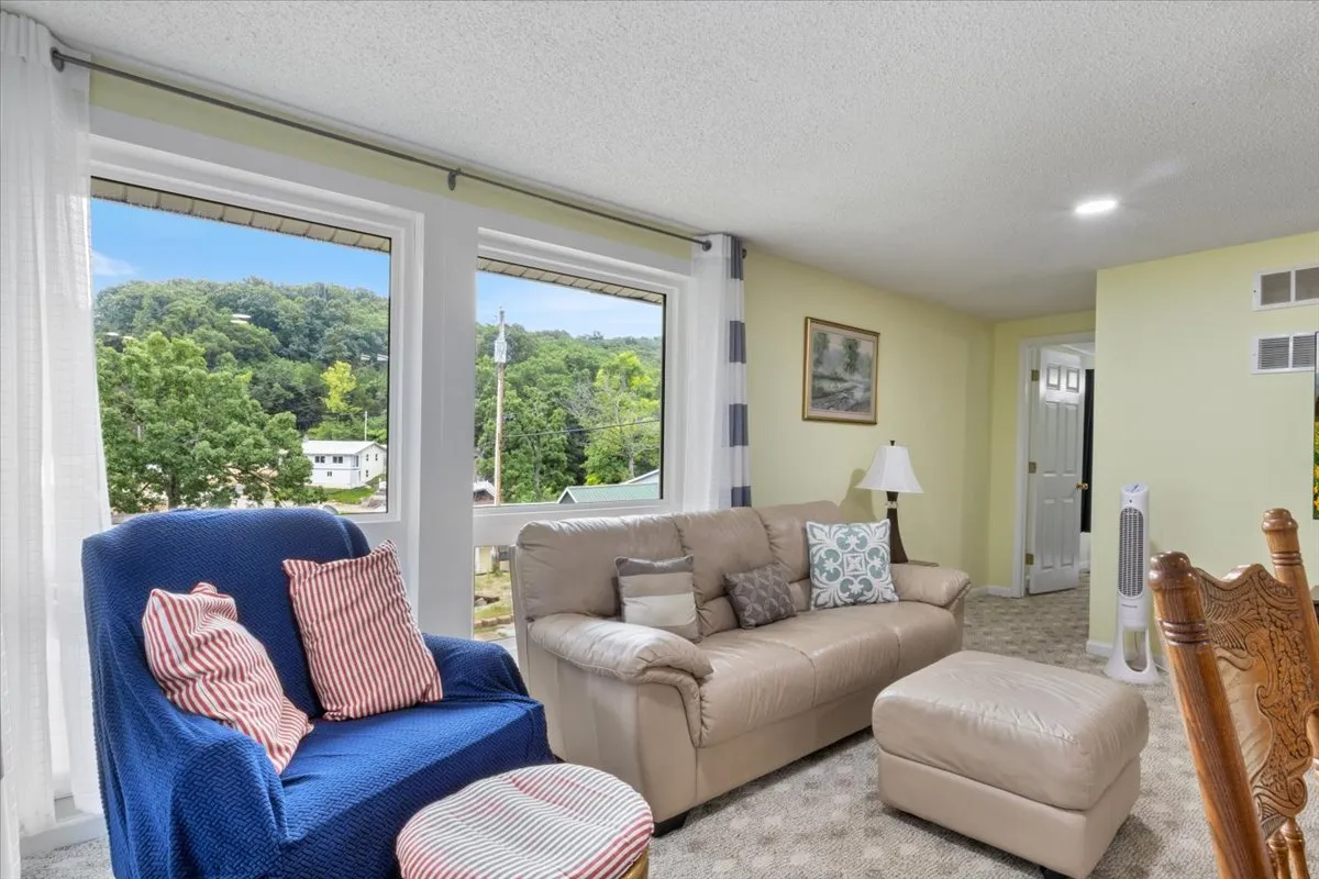 A cozy living room scene with a beige leather sofa, blue and white striped chair, and colorful throw pillows, all centered around large windows that bring in natural light and offer green views. The space radiates comfort and is clearly intended for relaxation, conversation, and enjoying the outdoors from within.