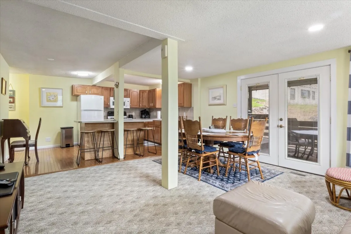 This image presents a broad view of the open living and dining area, showing a central dining table, visible kitchen, and a living space that flows seamlessly towards outdoor access. Large windows suggest abundant natural light and possible lake views, while the arrangement of furniture emphasizes togetherness and easy movement for group gatherings.