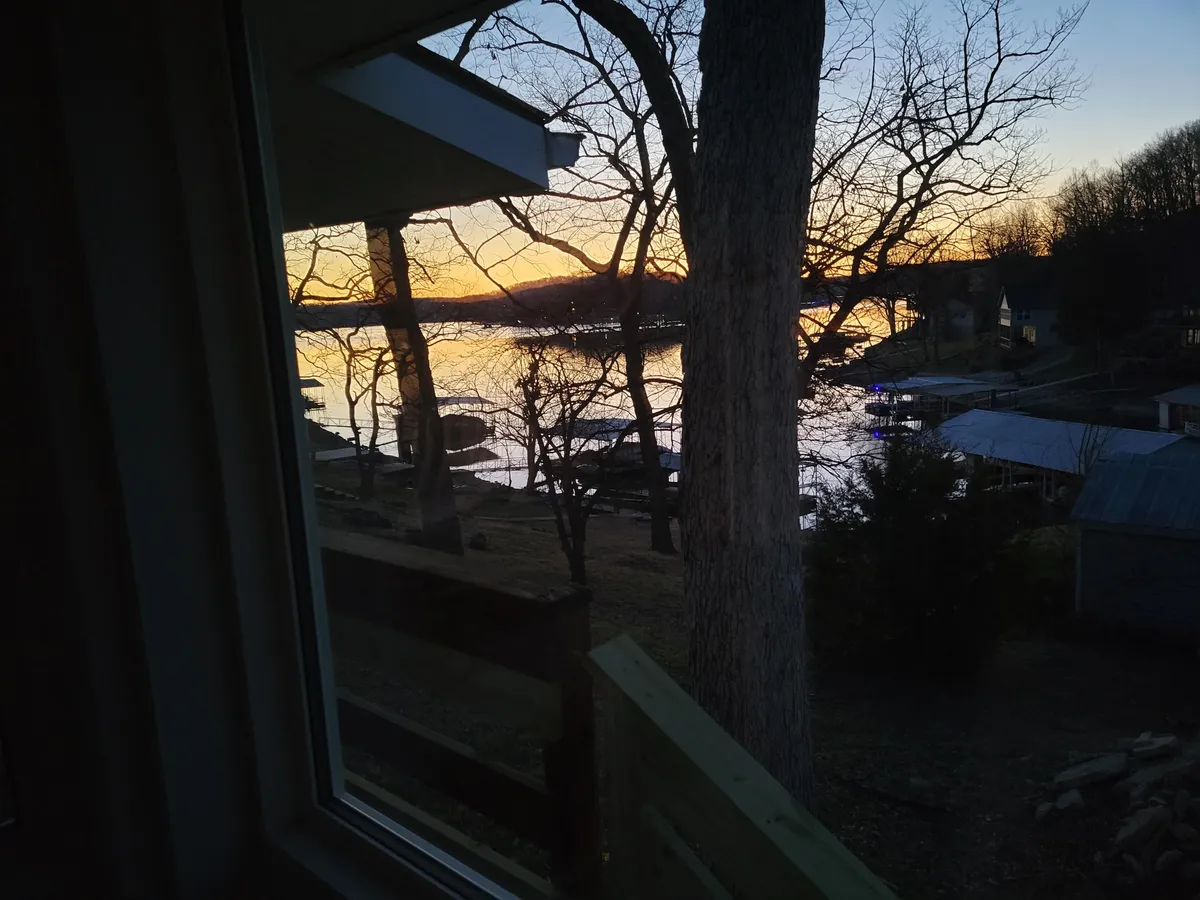 A stunning sunset over the lake, with reflections shimmering on the water, bare trees in the foreground, and distant houses nestled along the shore. The mood is tranquil and perfect for capturing the magic of an evening by the lake.